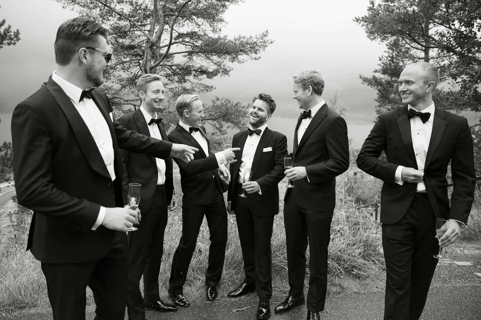 Gruppe menn i smoking som har en samtale utendørs i naturen, smilende og kledd for formell anledning.