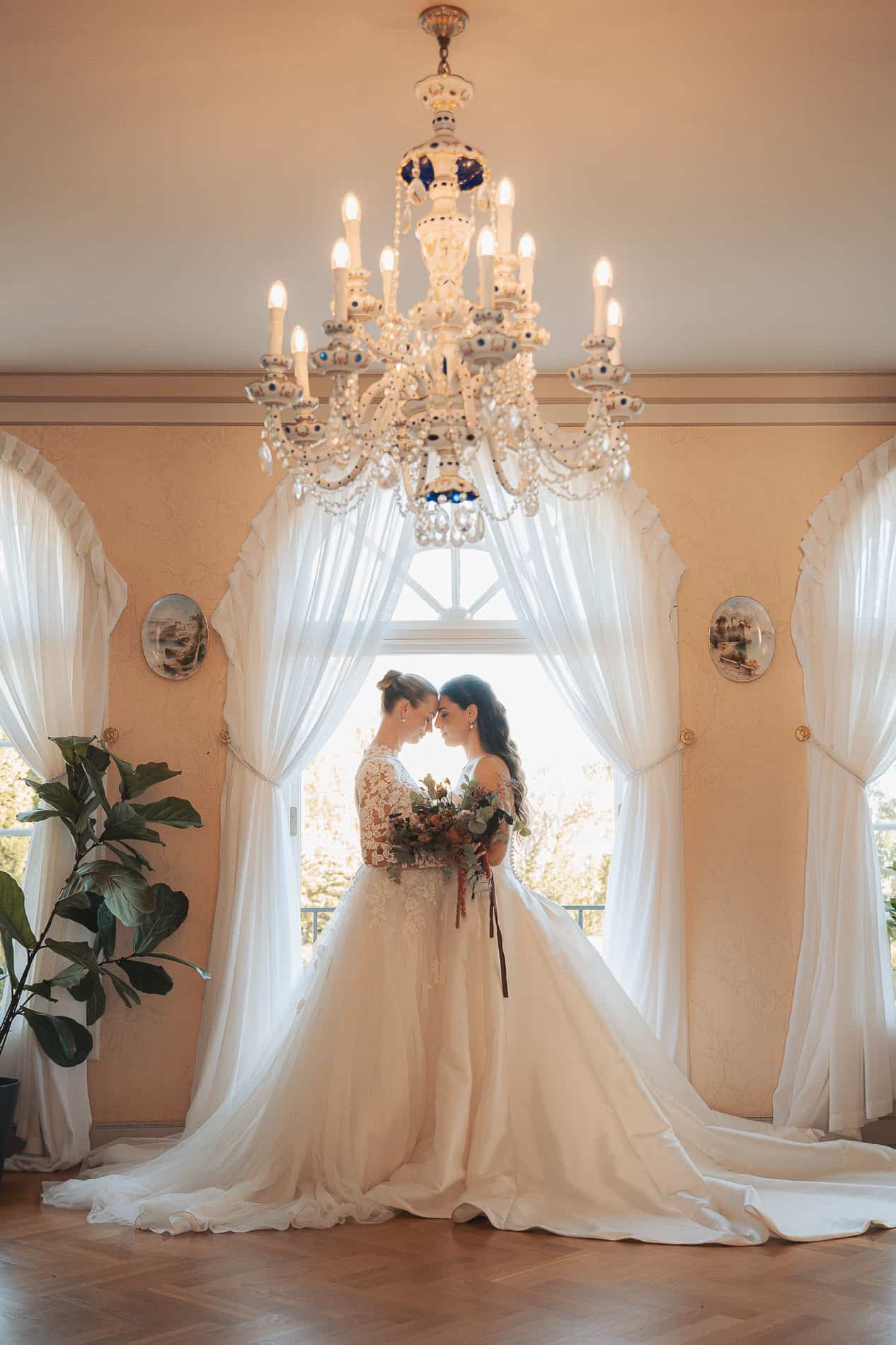 Bryllupsfoto av brudepar foran store vinduer med hvite gardiner, romantisk og elegant setting.