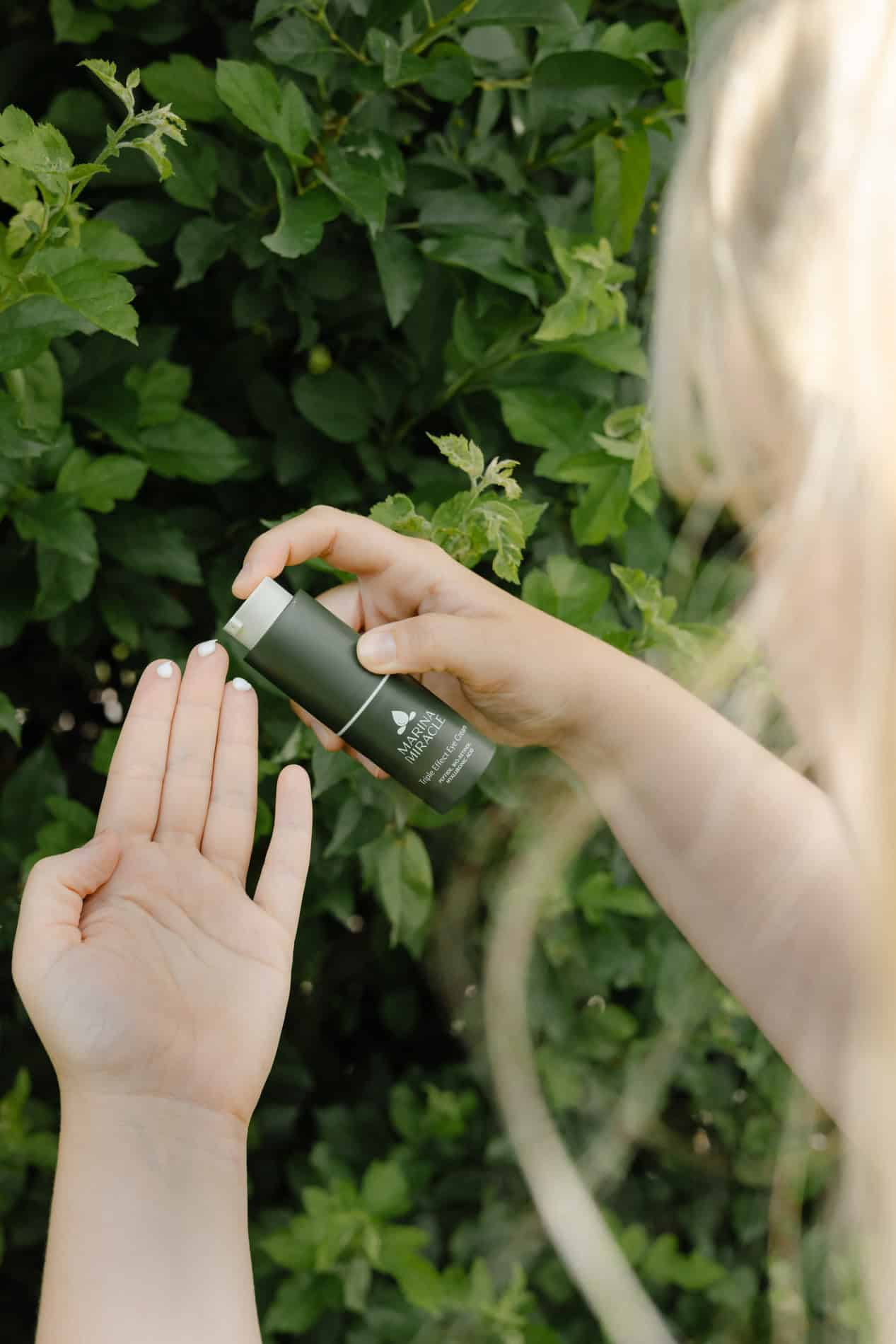 Person som påfører naturlig hudpleieprodukt på hånden foran grønt bladverk.