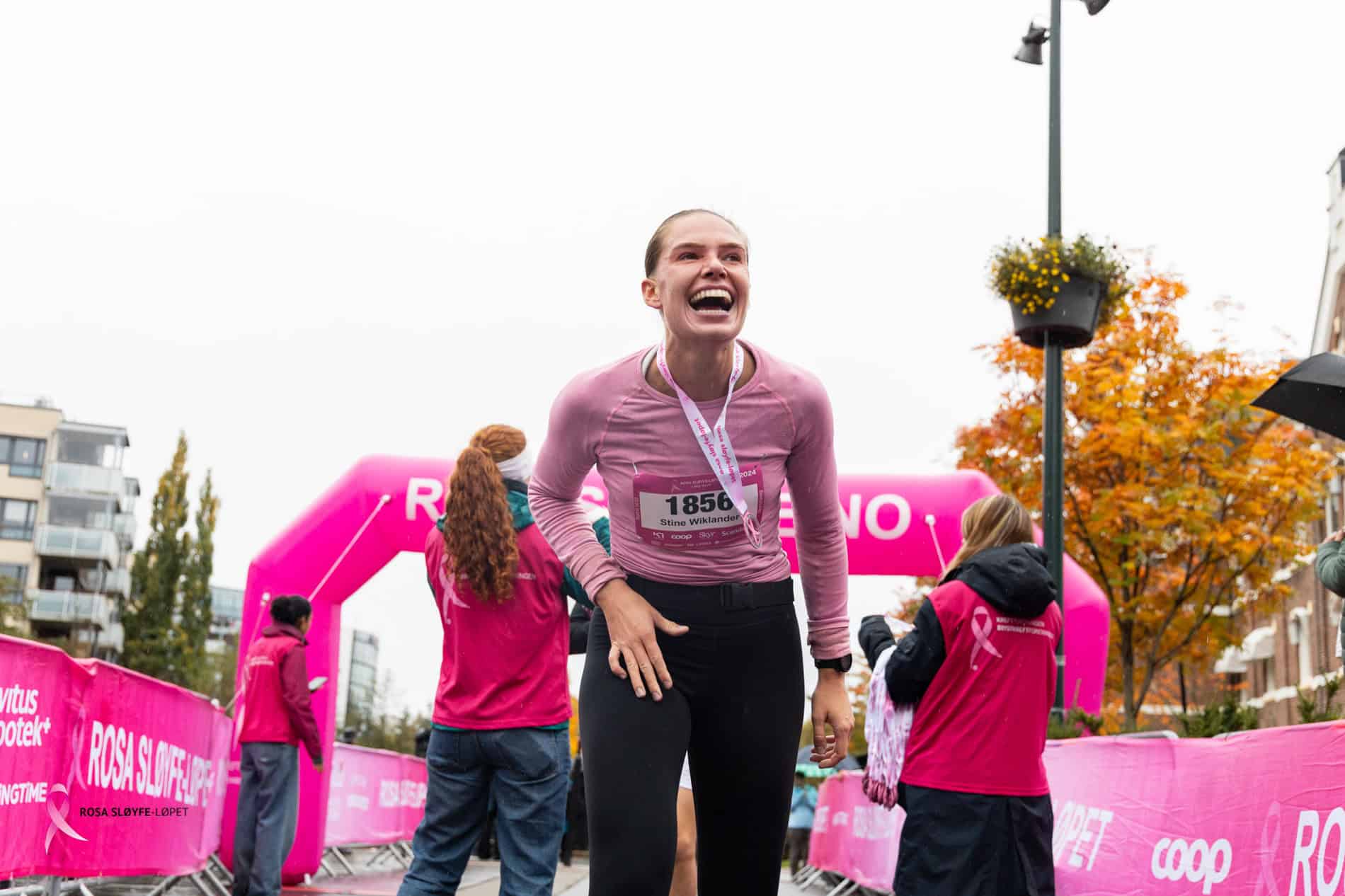 Løper smiler og jubler etter målgang under bedriftsløp, omgitt av supportere og fargerike arrangemen.