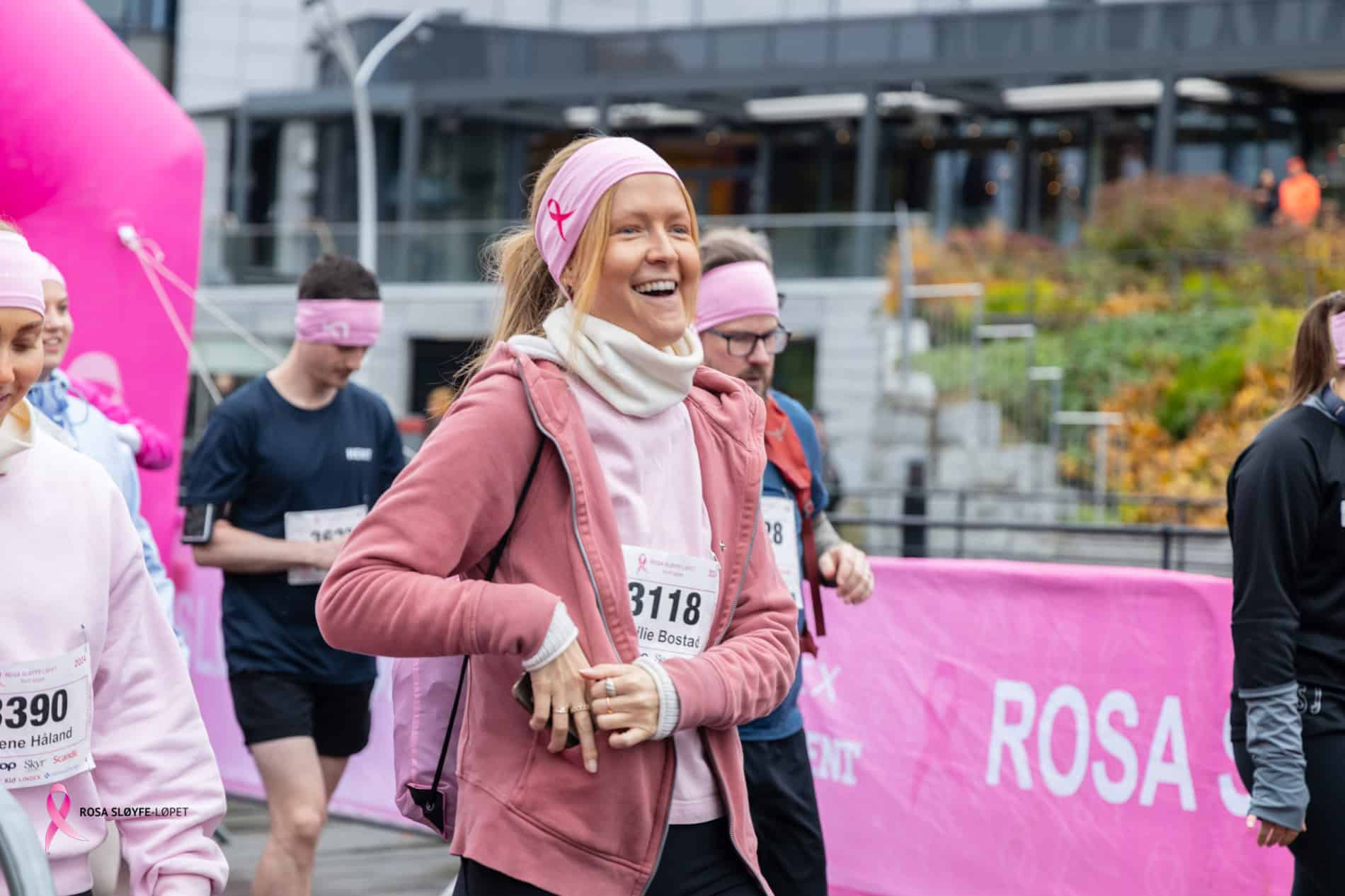 En kvinne smiler under bedriftsløp, omgitt av deltakere, med rosa dekor og moderne bygg i bakgrunnen.