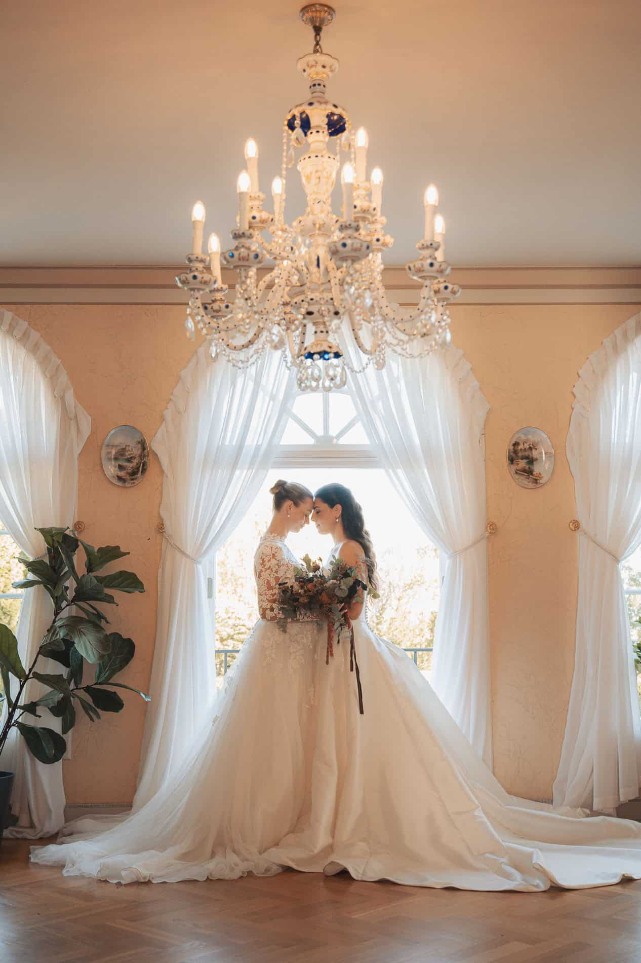 Bryllupsfoto av brudepar under elegant lysekrone i innendørs setting.