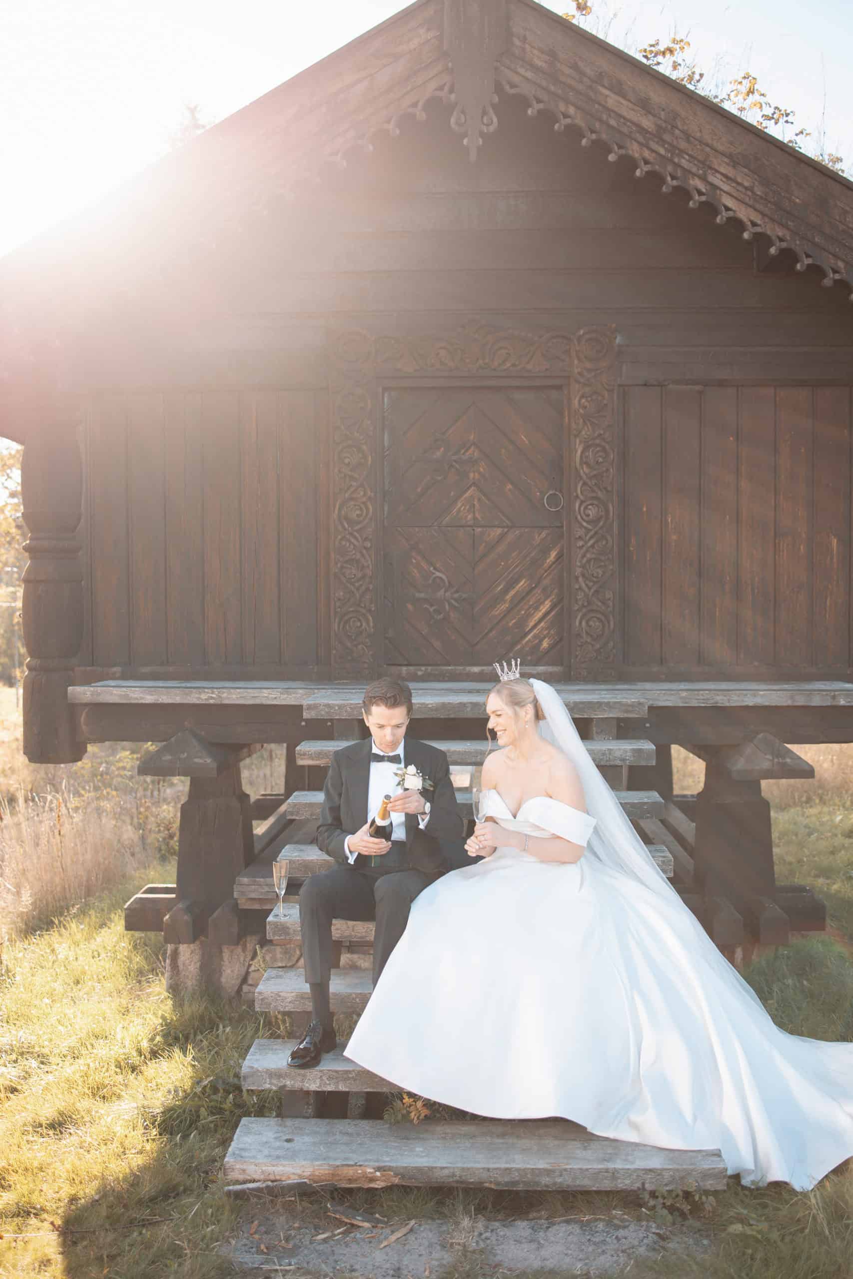 Brudepar i bryllupsantrekk foran et tradisjonelt trehus, sollys i bakgrunnen.