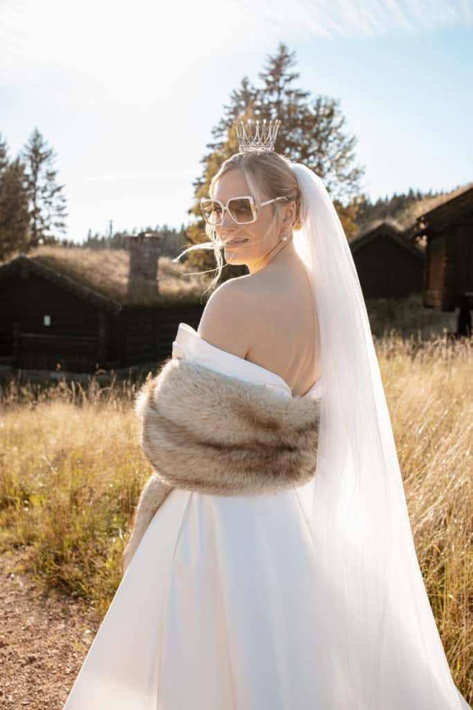 Brud i vakker hvit kjole med pels og solbriller, fotografert utendørs i naturlig lys.