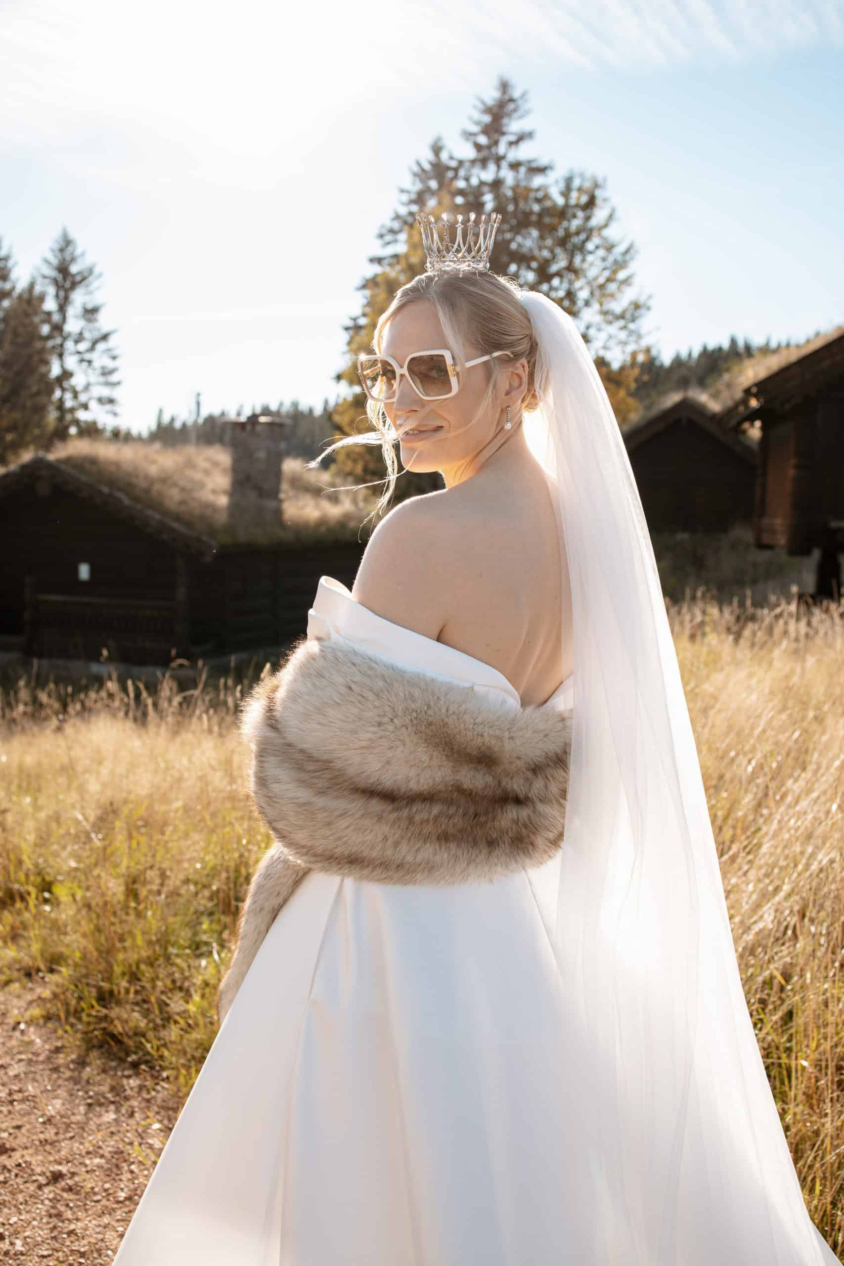 Brud i vakker hvit kjole med pels og solbriller, fotografert utendørs i naturlig lys.