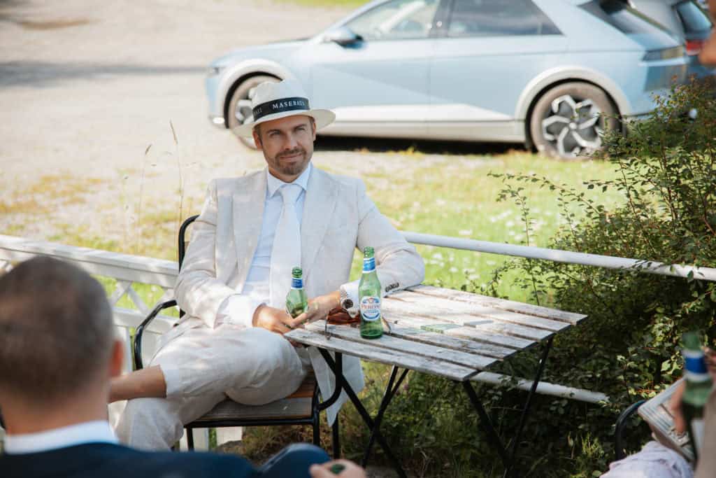 Mann i lys dress med hat nyter utendørsmiddag med venner, bil i bakgrunnen.