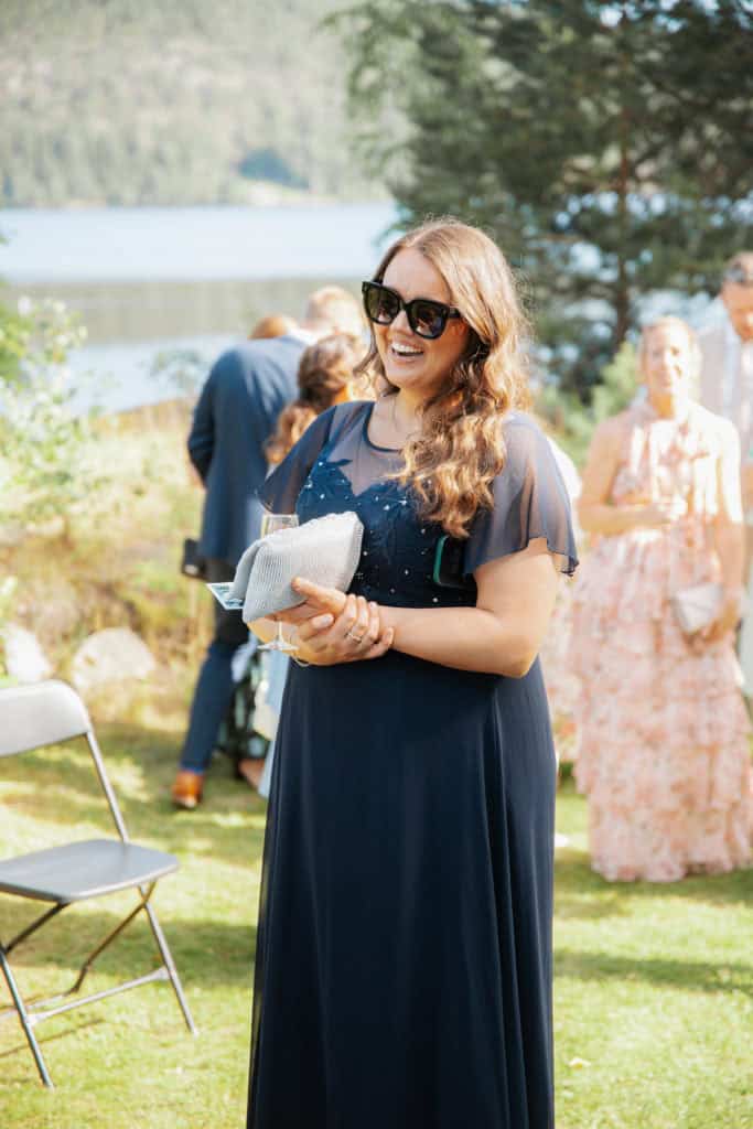 Kvinne i elegant blå kjole med solbriller, smilende under utendørs fotoshoot.