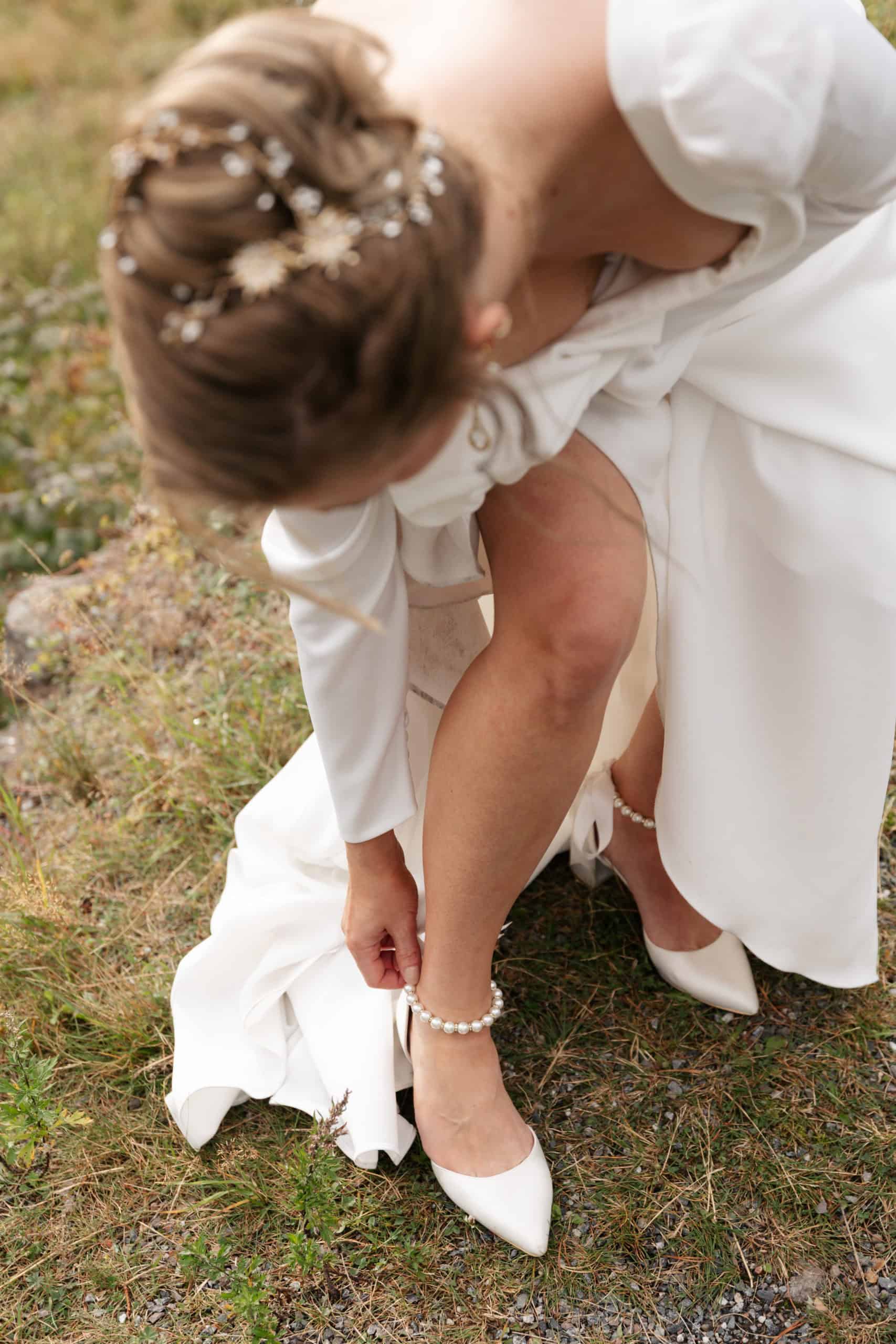 Brud som justerer skoene sine på gress, fokus på hvit kjole og blomster i håret.