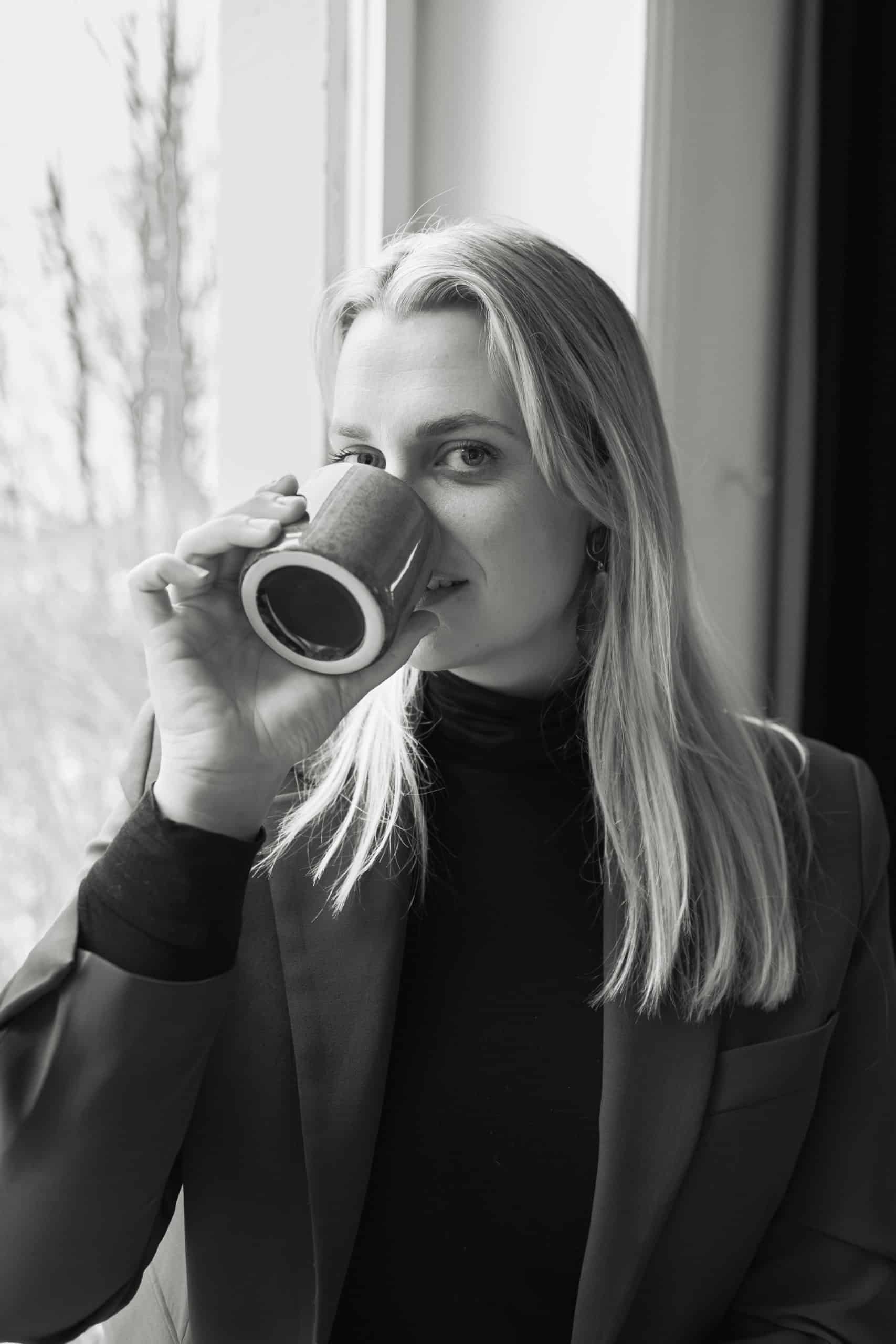 Heidi Henriksen nyter en kopp kaffe foran et vindu, i svart-hvitt bilde.